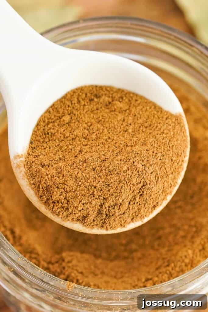 Close-up of a heaping spoonful of freshly mixed apple pie spice being poured into a small glass jar, highlighting the texture and richness of the blend