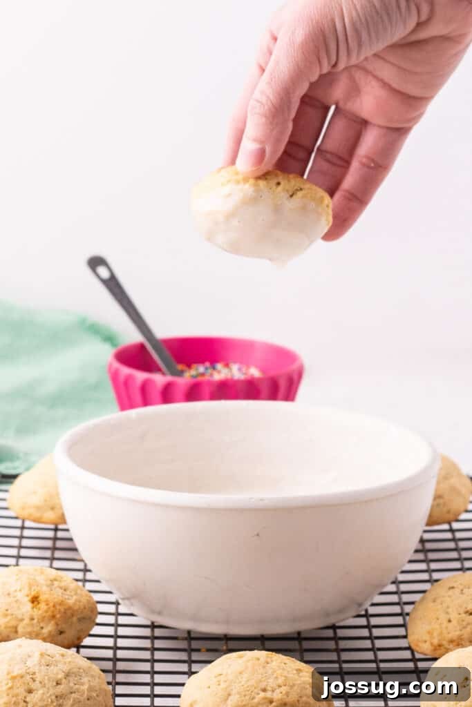 Pillowy Italian Ricotta Cookies 14 Hand dipping a ricotta cookie into almond glaze with a bowl of sprinkles in the background