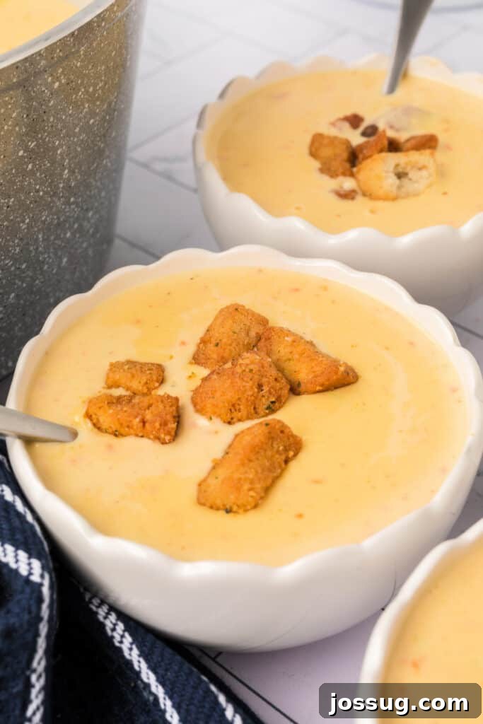 A close-up of a white scalloped bowl filled with creamy cheese soup, topped with golden croutons and served with a spoon. Steam gently rises from the hot soup, emphasizing its warmth and inviting texture.