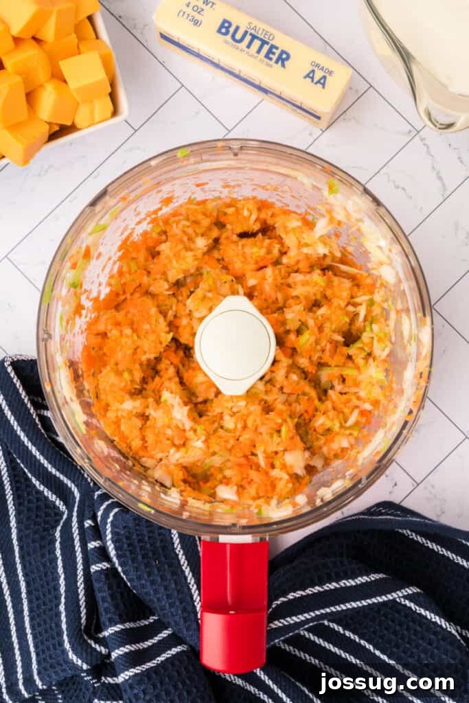 Finely chopped carrots, onion, and celery are blended together in a food processor bowl, sitting on a navy striped kitchen towel, ready to be added to the pot.