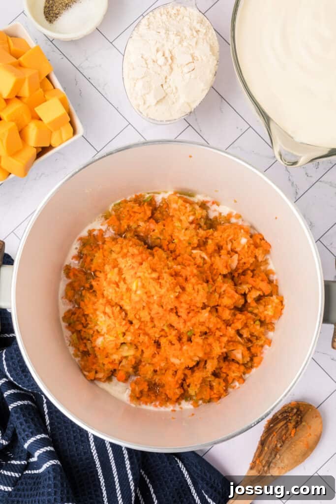 The vibrant pureed carrots, onion, and celery are now added to the melted butter in the large white pot. Nearby, other key ingredients like flour, cubed Velveeta cheese, and heavy cream await their turn, showcasing the sequential steps of the recipe.