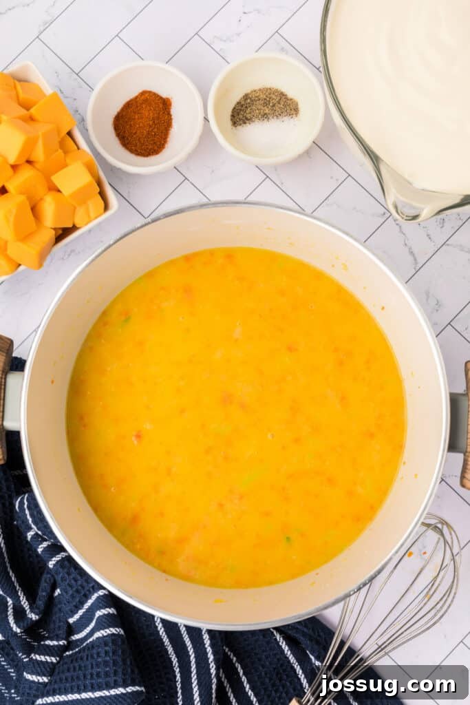 Vegetable broth has been gradually whisked into the rich vegetable roux, transforming it into a smooth, golden soup base. A whisk rests on the side of the pot, indicating the ongoing blending process.