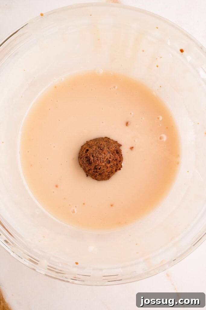 Fried donut hole dipped into vanilla glaze in a glass bowl