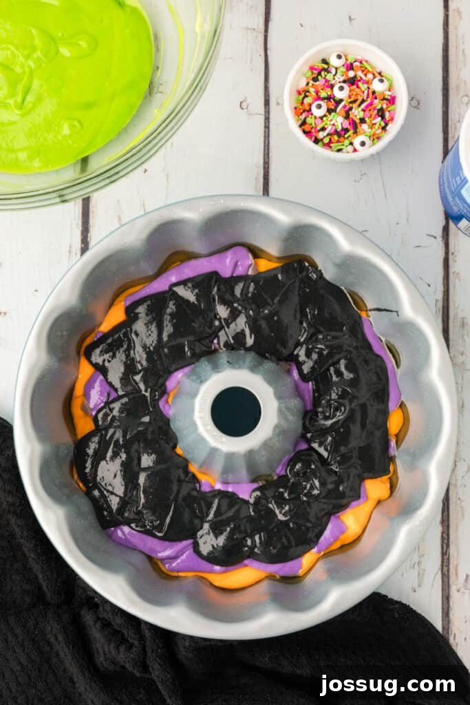 Bewitched Bundt Cake 13 Bundt pan with layered orange, purple, and black cake batter, with a bowl of green batter and Halloween sprinkles nearby.