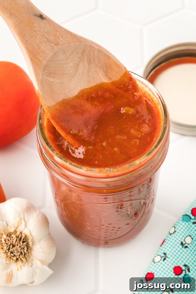 wooden spoon scooping homemade enchilada sauce out of a glass jar, showing its rich color and inviting texture