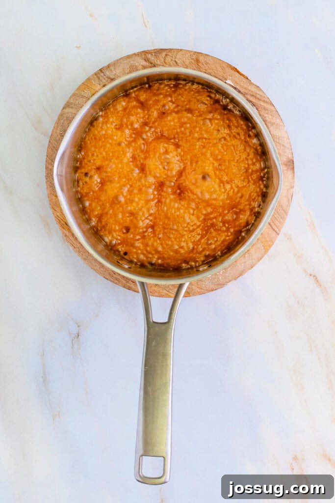 Saucepan filled with a shimmering, actively bubbling golden caramel mixture, just removed from the heat and resting on a wooden trivet, signaling its readiness for the next crucial step of adding vanilla and baking soda.