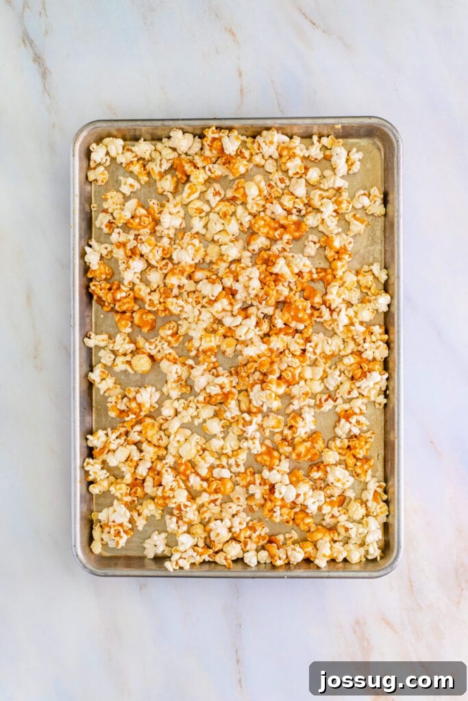 Baking sheet showcasing plain popcorn partially coated with a rich, golden caramel sauce, appearing somewhat clumpy but awaiting further baking in the oven to achieve a full, even, and crispy glaze.
