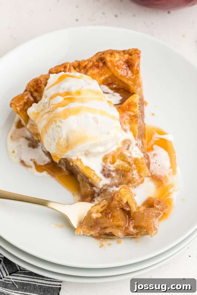 Slice of apple pie with melting ice cream and caramel drizzle on a plate, showcasing a golden crust and rich apple filling.