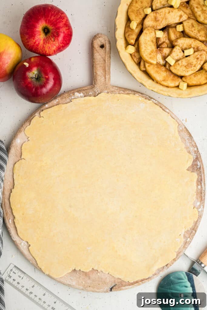 A sheet of perfectly rolled out pie dough rests on a wooden board, positioned thoughtfully next to the already filled apple pie crust, ready for its lattice top.