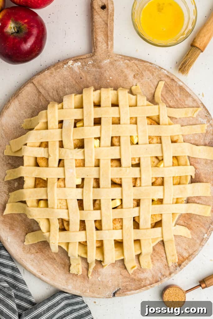An unbaked lattice top apple pie, with its edges awaiting trimming, alongside a small bowl of egg wash and a sprinkle of coarse sugar, poised for baking.