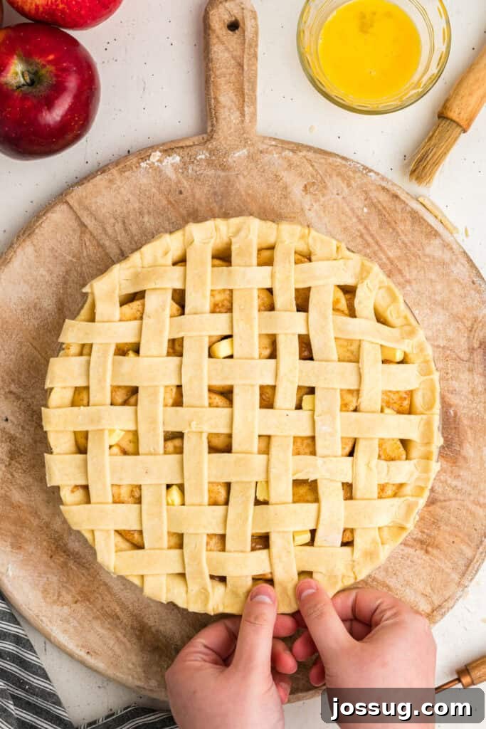 Hands delicately folding the trimmed lattice edges underneath the main pie crust to create a clean, crimped, and aesthetically pleasing border for the apple pie.