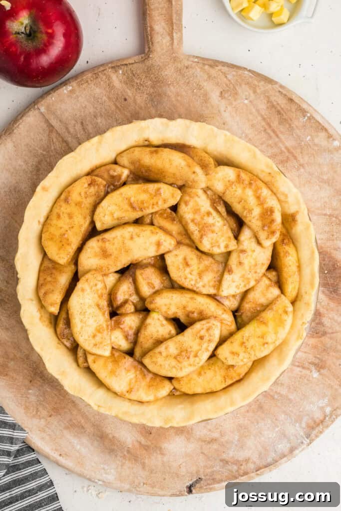An unbaked, rustic pie crust nestled in its pan, generously filled with a mound of seasoned apple slices, resting on a wooden board.