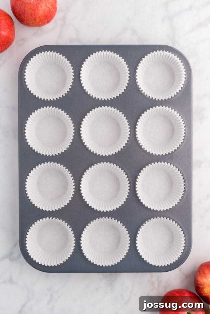 Cider Mill Muffins 4 Muffin pan lined with white paper liners surrounded by fresh apples on a marble surface.