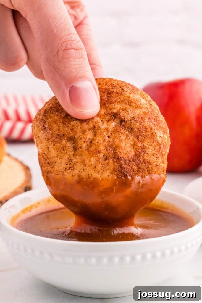 A close-up shot of an individual apple pie bomb being luxuriantly dipped into warm, glossy spiced caramel sauce.