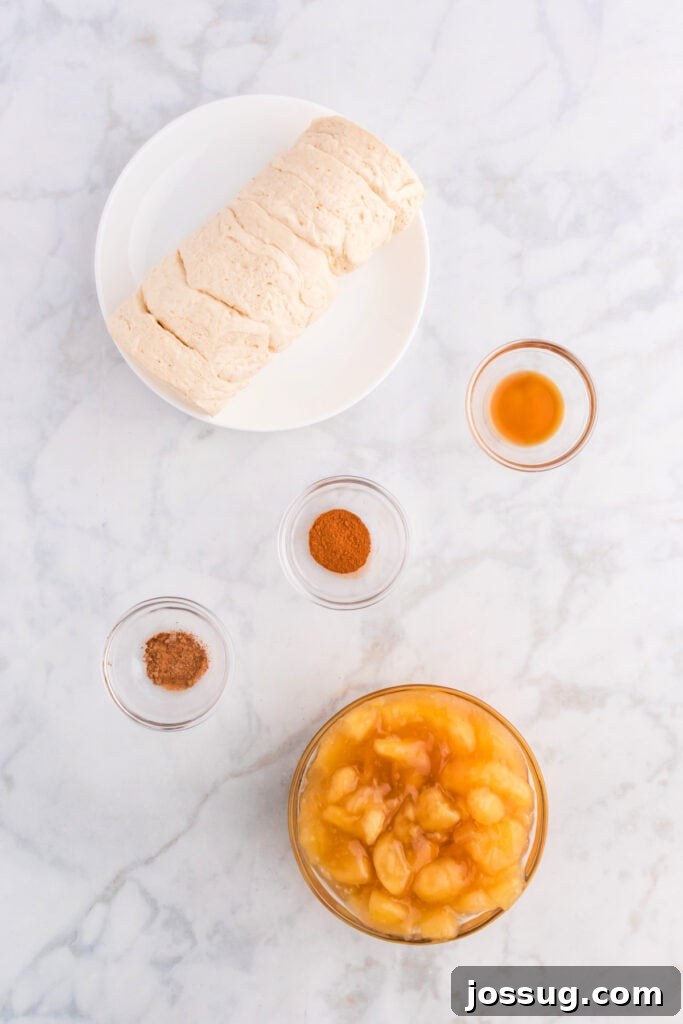Ingredients for apple pie bombs including biscuit dough, apple pie filling, vanilla, and spices neatly arranged on a marble counter.