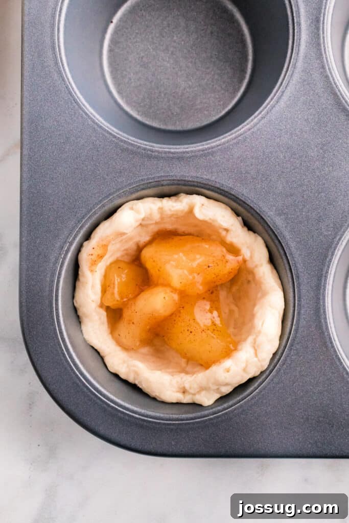 A biscuit dough cup generously filled with spiced apple pie filling, nestled in a muffin tin cavity.