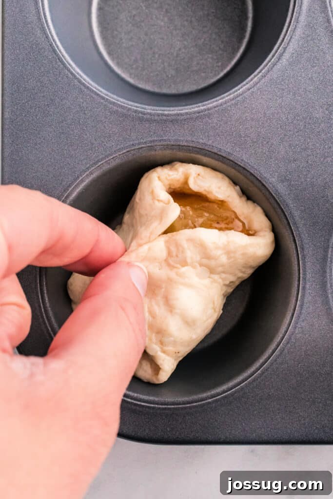 Hands carefully folding biscuit dough over apple pie filling, meticulously pinching to form a sealed apple pie bomb.