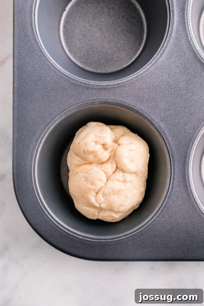 A perfectly sealed apple pie bomb dough ball resting in a muffin pan cavity, awaiting its turn in the oven.