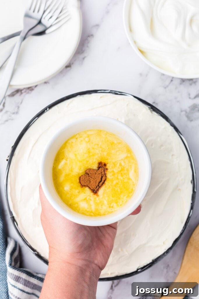 Decadent No-Bake RumChata Pumpkin Cheesecake 16 Hand holding a small bowl of melted butter and cinnamon mixture above the cheesecake, ready to make the cinnamon drizzle topping.