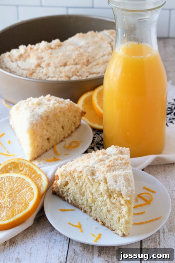 A generous slice of Orange Streusel Coffee Cake on a white plate, with a pitcher of fresh orange juice in the background.