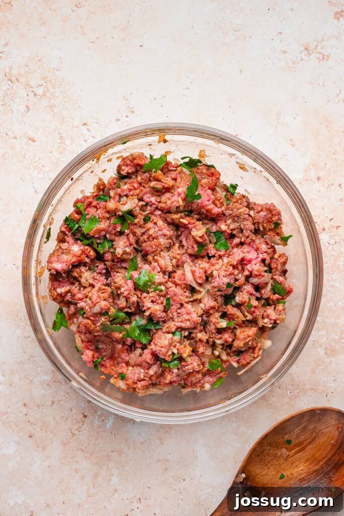 Arayes filling mixture of ground beef, lamb, parsley, onion, garlic, and spices thoroughly mixed together in a glass bowl with a wooden spoon beside it.