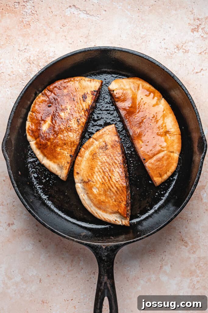 Three stuffed pitas flipped in a cast iron skillet, golden brown and crisping on the surface as they cook for arayes.