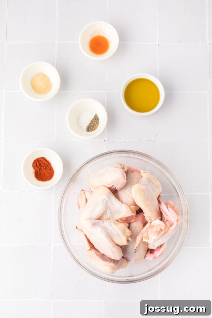 Smoky Sweet Air Fryer Chipotle Honey Glazed Wings 3 Raw chicken wings in a glass bowl with small bowls of olive oil, hot sauce, garlic powder, paprika, salt, and pepper on a white tile surface.