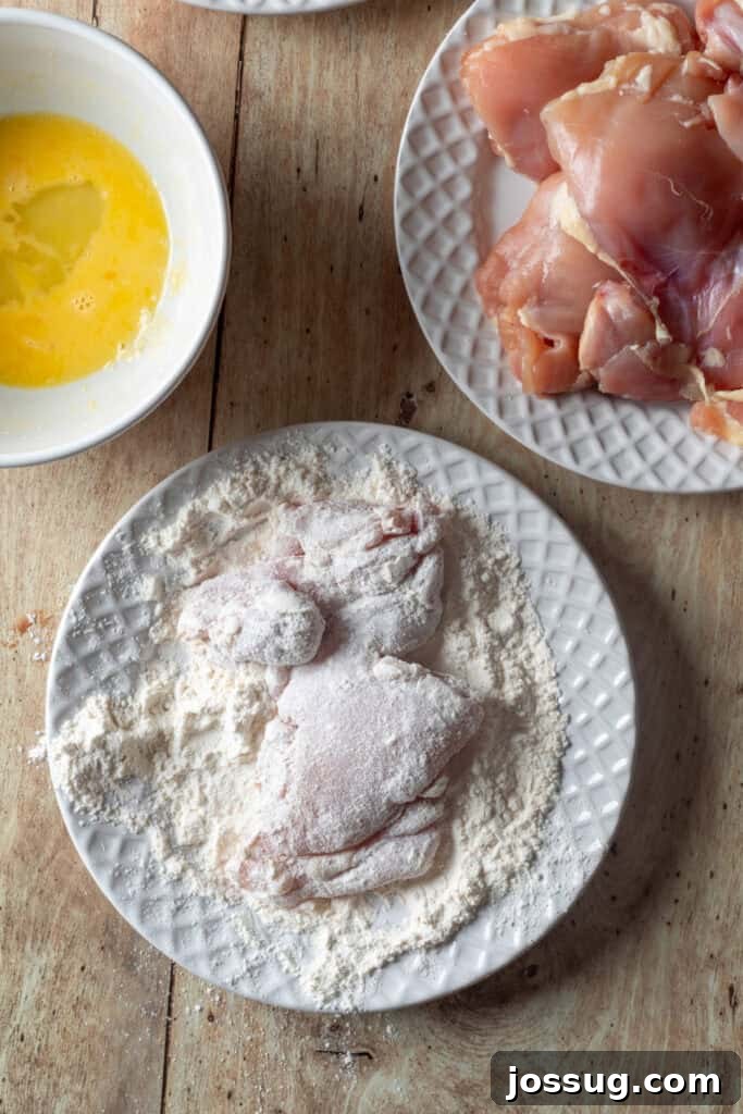 Cowboy Butter Chicken Crunch Sandwich 6 Chicken thighs dredged in all-purpose flour on a white plate, with bowls of beaten eggs and raw chicken waiting to be breaded.