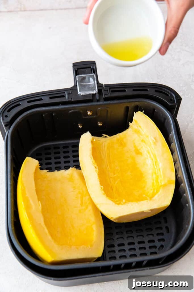 Hand holding a small bowl of oil over two spaghetti squash halves in the air fryer basket.