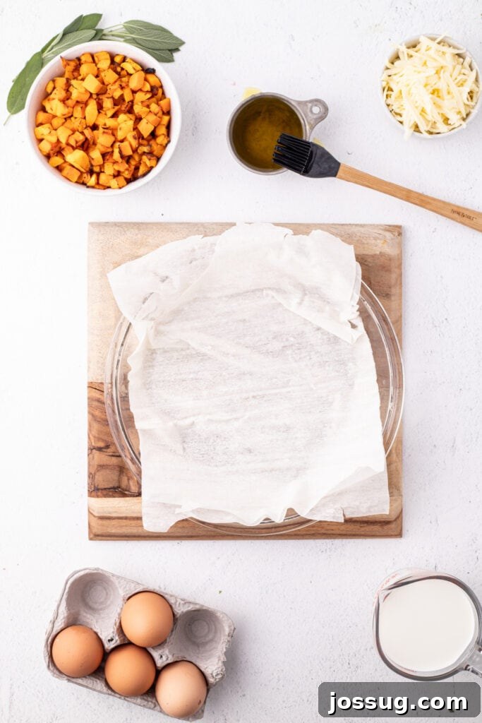 A pie dish lined with the first sheet of phyllo dough on a wooden board surrounded by bowls of squash, shredded cheese, sage leaves, eggs, olive oil, and milk.