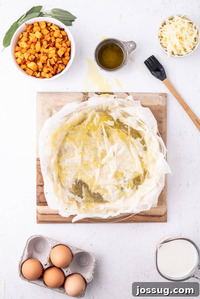 Multiple layers of phyllo dough brushed with olive oil in a pie dish, surrounded by roasted squash, shredded cheese, eggs, and milk.
