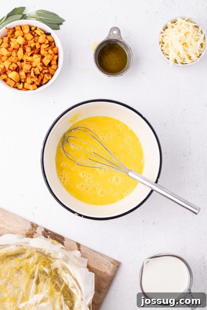 Mixing bowl with beaten eggs and whisk, with nearby bowls of roasted squash, shredded cheese, olive oil, and a prepared phyllo crust.