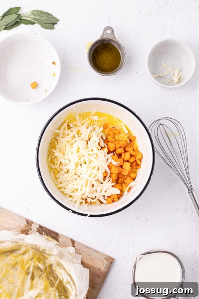 Mixing bowl filled with beaten eggs, roasted squash, and shredded Gruyère cheese, with whisk and prepared crust nearby.