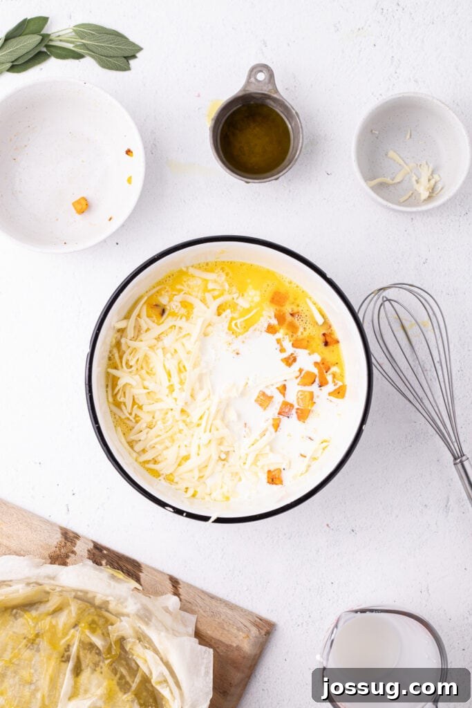 Egg mixture in bowl with added half and half, roasted squash, and shredded Gruyère cheese, with whisk and phyllo crust nearby.
