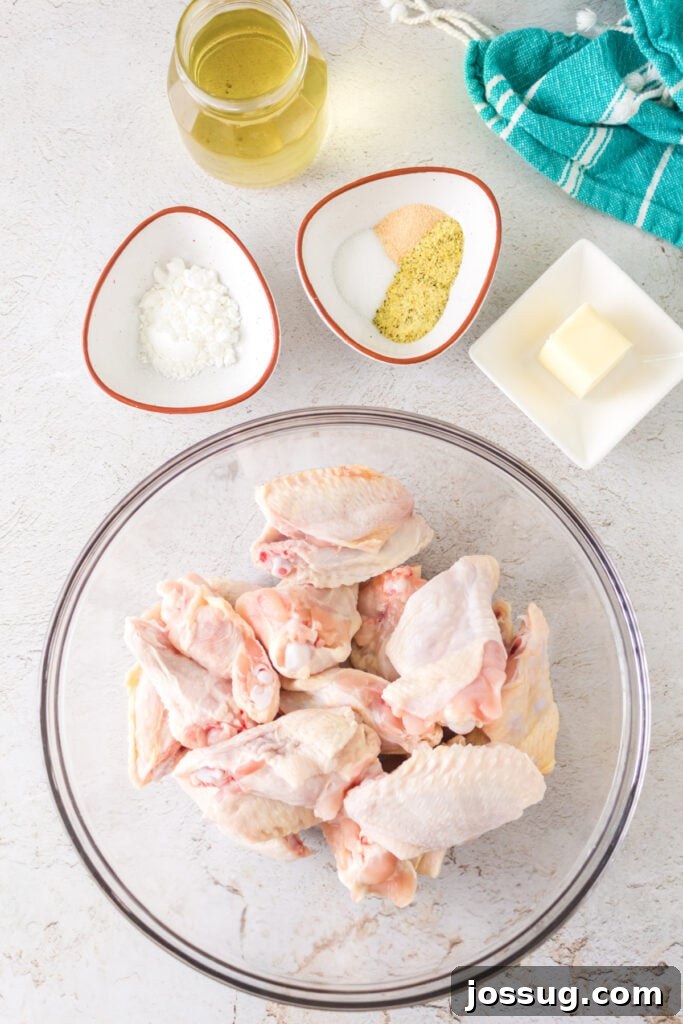 Overhead shot of raw chicken wings in a glass bowl with small bowls of cornstarch, lemon pepper seasoning, melted butter, and olive oil on the side, ready for preparation.