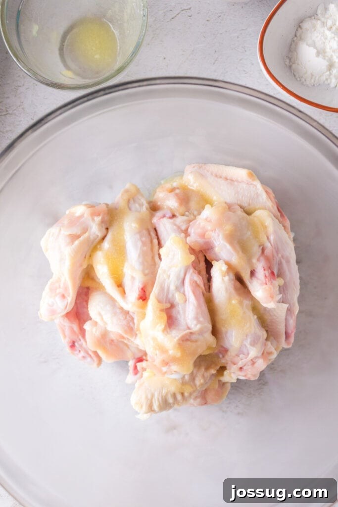 Raw chicken wings in a clear mixing bowl, being drizzled with melted butter for even coating.