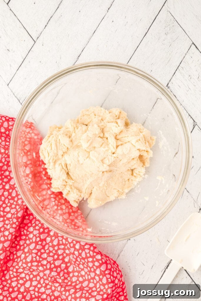 A bowl of freshly mixed shortcake dough, ready to be rolled out, next to a red patterned kitchen towel and a spatula.