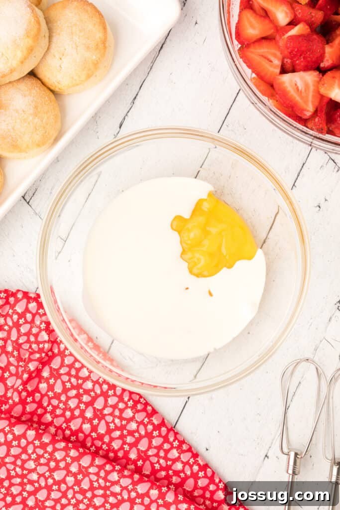 A glass bowl containing heavy cream, with a dollop of bright yellow lemon curd waiting to be mixed in, alongside fresh strawberries and baked shortcakes.