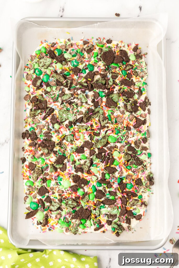 A large, rustic slab of homemade mint chocolate bark, expertly studded with vibrant green M&M's, chunky crushed mint Oreos, and a shower of rainbow sprinkles, resting on a flat surface, ready to be broken into individual pieces.