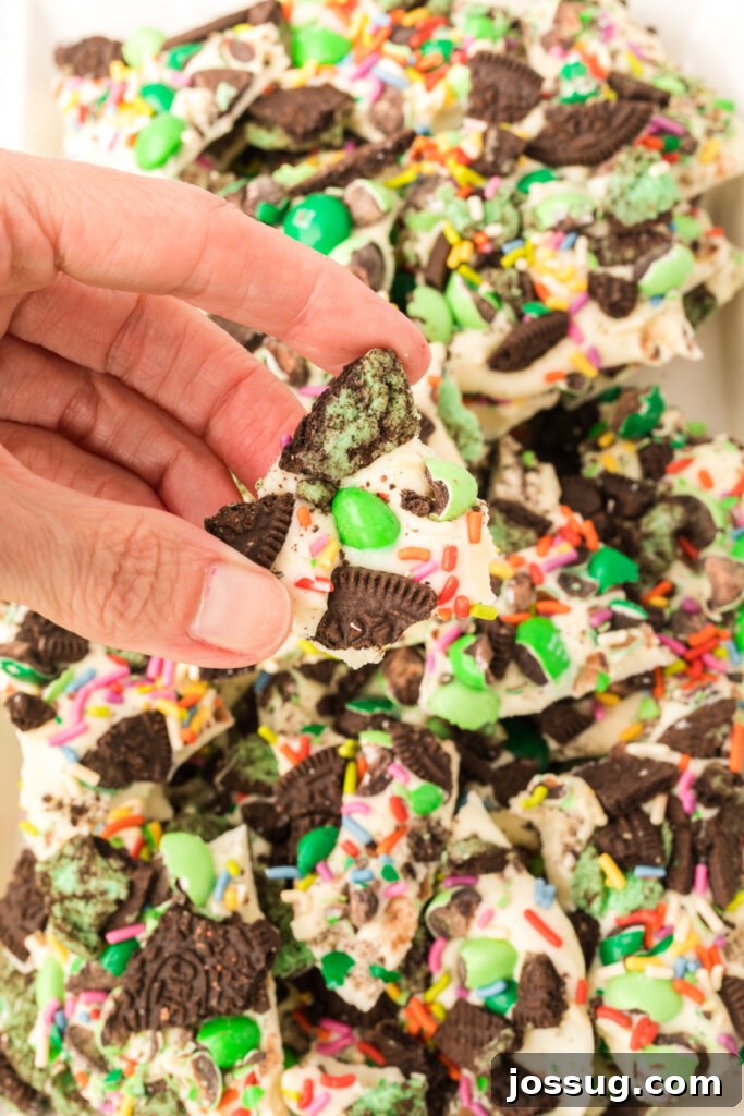 A hand gently holds a freshly broken piece of thick Loaded Leprechaun Mint Bark, showcasing the delightful layers of mint candy, green M&M's, crushed mint Oreos, and rainbow sprinkles embedded within.