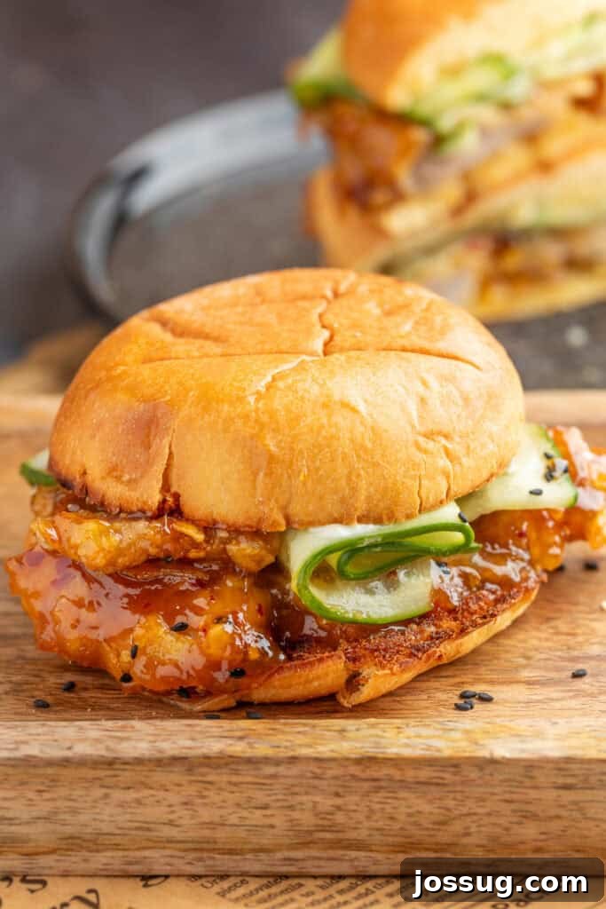 Close-up of a finished orange chicken sandwich with sauced chicken and crisp cucumber ribbons on a toasted brioche bun, sprinkled with sesame seeds.