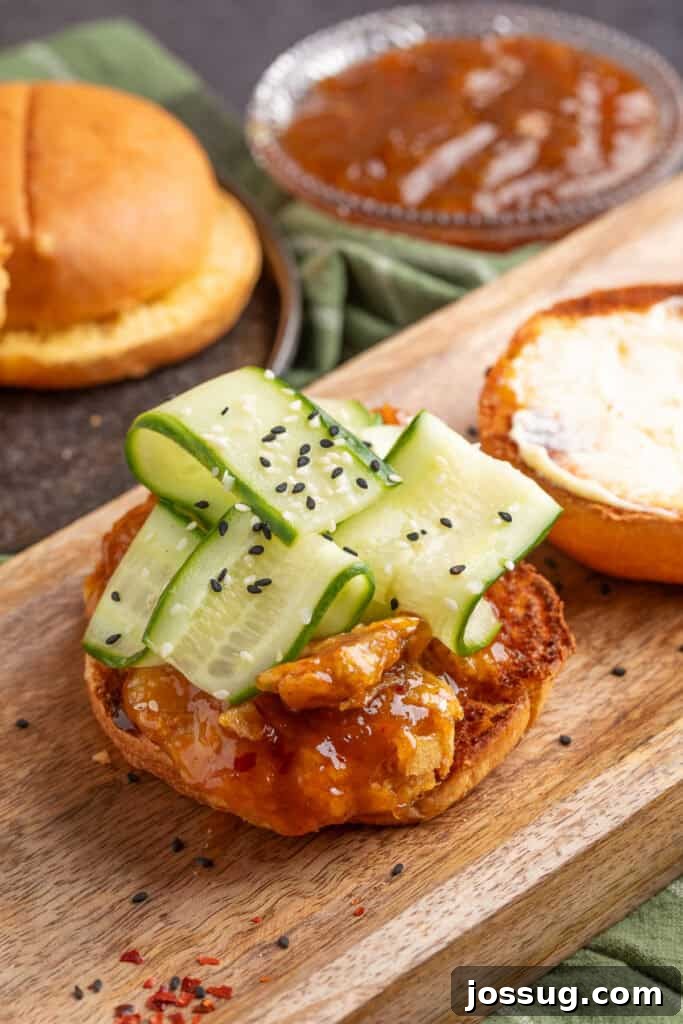 Open sandwich on a wooden board with sauced chicken, mayonnaise on the bun, refreshing cucumber ribbons, and sprinkled sesame seeds.