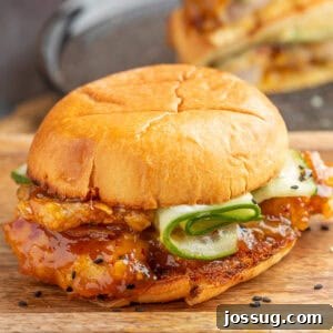 Square image of an orange chicken sandwich with saucy chicken, cucumber ribbons, and sesame seeds on a toasted bun.
