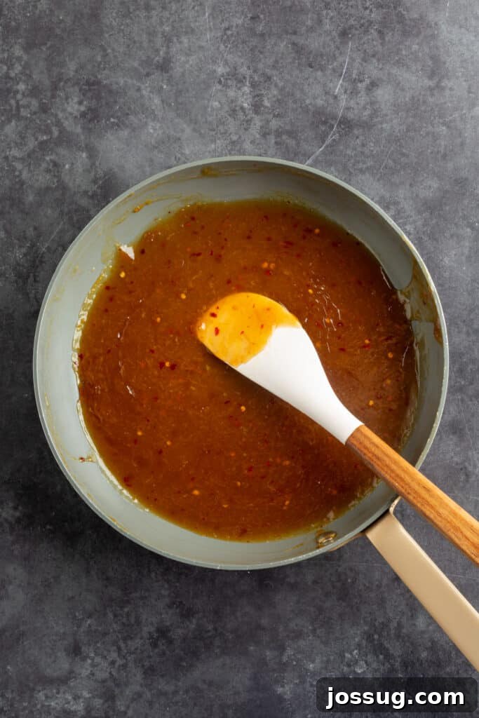 Saucepan filled with homemade orange sauce simmering, visible red pepper flakes, stirred with a white spatula.