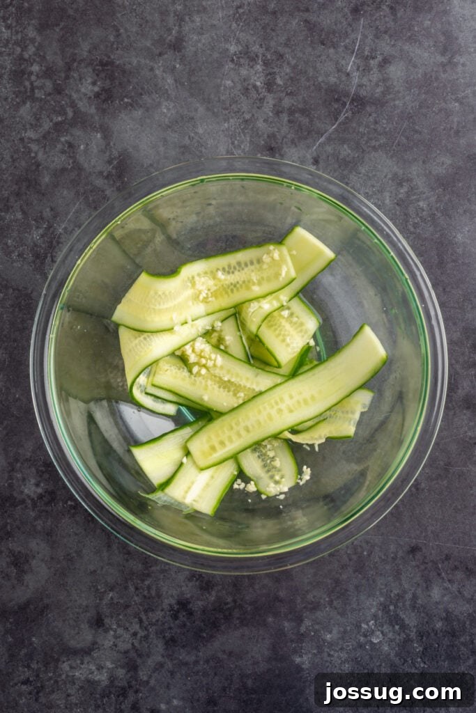 Glass bowl of thinly sliced cucumber ribbons tossed with minced garlic and rice vinegar for a quick cucumber salad.