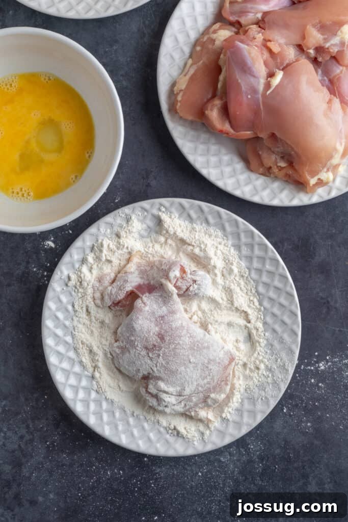 Raw chicken thigh coated in flour on a white plate next to beaten egg wash and uncooked chicken thighs, prepared for breading.