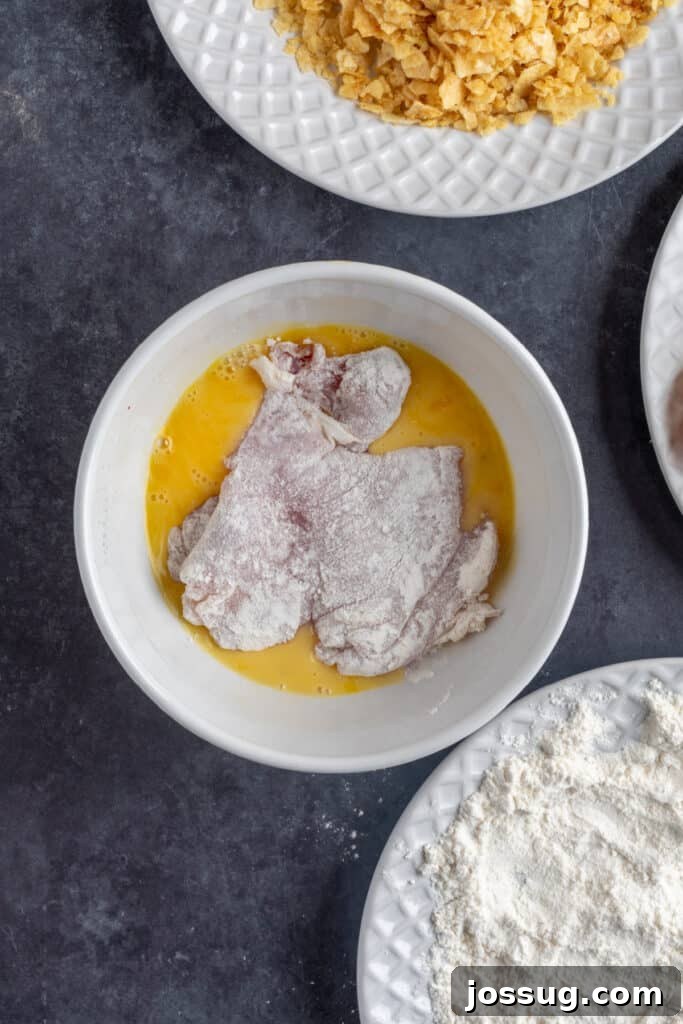 Chicken thigh dipped into beaten egg wash after dredging in flour, ready for coating with crushed chips.
