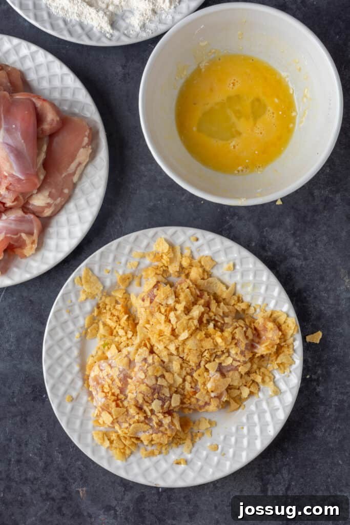 Chicken thigh coated in crushed potato chips, sitting on a plate beside bowls of egg wash and flour, showing the breading stages.