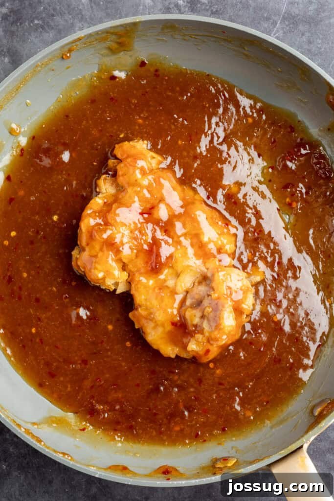 Breaded air-fried chicken thigh being coated in sticky homemade orange sauce inside a skillet, ready for assembly.