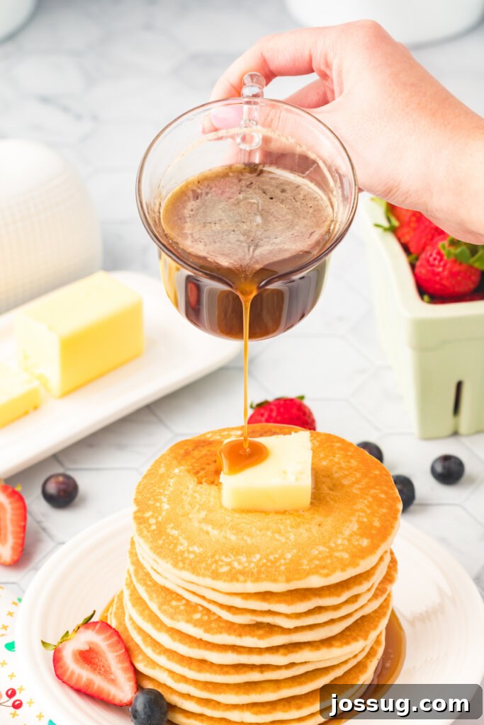 Golden Vanilla Pancake Nectar 4 A hand gently pouring luscious homemade vanilla pancake syrup over a towering stack of golden pancakes, creating an inviting visual.